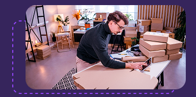 Person packing a box on a desk surrounded by stacks of parcels in a home office setting.