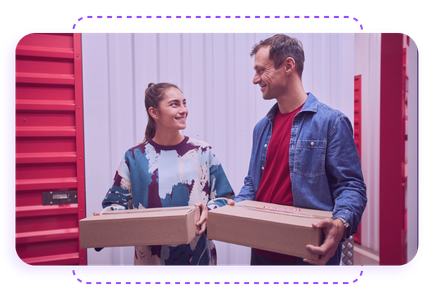 Two people smiling at each other while holding cardboard boxes in a storage facility with red and white walls.
