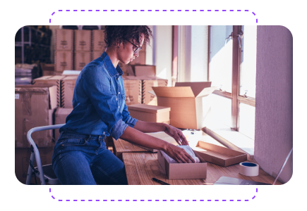 A person in a denim shirt sitting at a desk, packing items into a cardboard box in a room filled with stacked boxes.