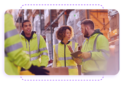 Workers in high-visibility jackets discussing logistics in a warehouse environment, surrounded by racks of packages.