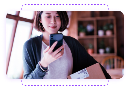 Woman holding a phone and a package with a label, standing indoors near a window with shelves in the background.