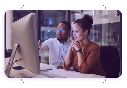 Two people sit at a desk, focused on a computer screen. One points at the monitor while the other listens attentively.