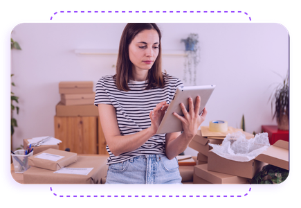 A woman in a striped shirt uses a tablet amidst a room filled with cardboard boxes and packing materials.