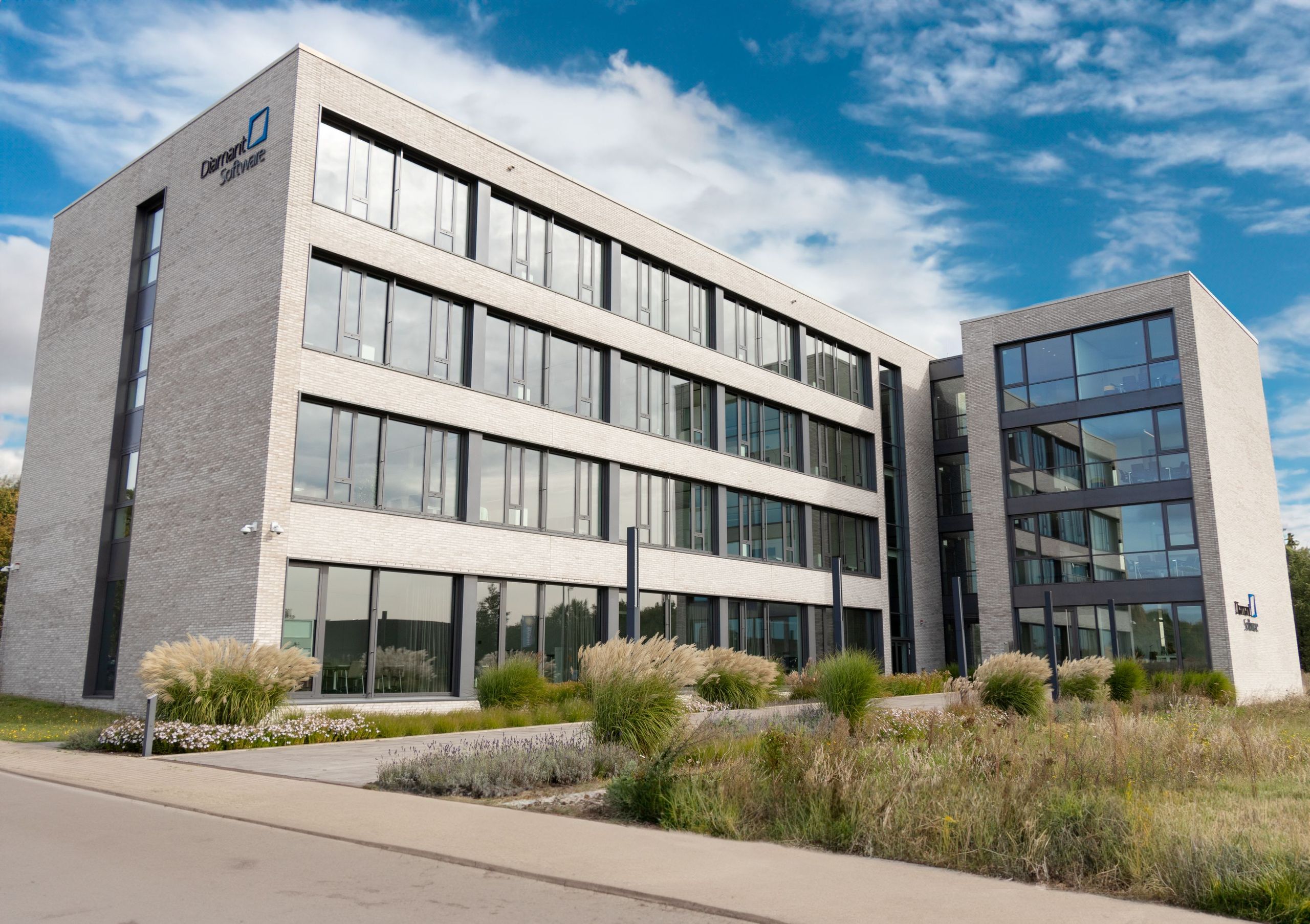 Modern four-story office building with large windows, surrounded by landscaped greenery against a partly cloudy sky.