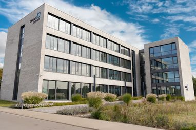 Modern four-story office building with large windows, surrounded by landscaped greenery against a partly cloudy sky.