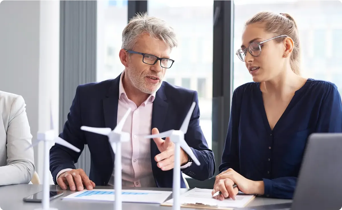 Two professionals discussing wind energy models at a table, with small wind turbine models and documents in front of them.