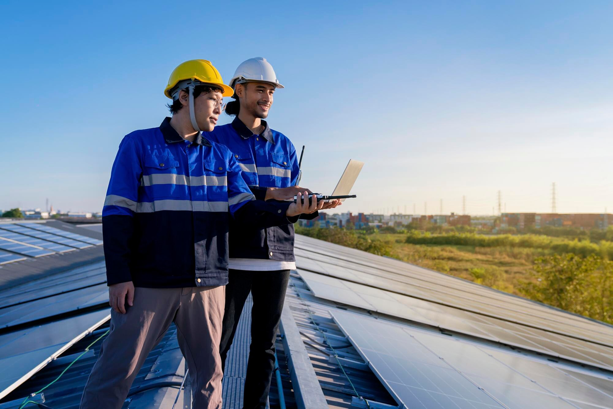 Two workers in blue jackets and helmets stand on a rooftop with solar panels, using a laptop, overlooking a landscape.