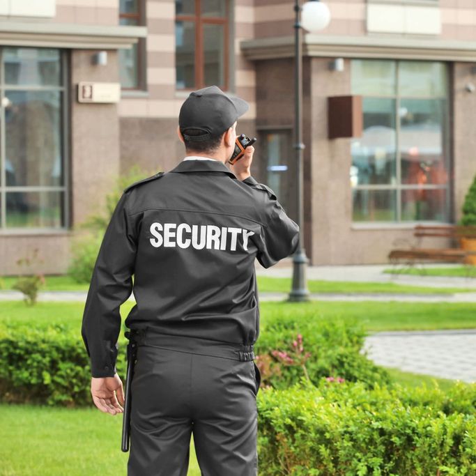 Security guard in uniform with "SECURITY" on the back, holding a walkie-talkie, stands in a landscaped area near a building.