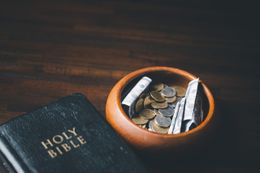 A wooden bowl filled with coins and rolled bills next to a Holy Bible on a wooden surface.