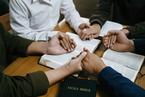 Group of people holding hands in prayer around a table with open Bibles and a Holy Bible in the center, representing the type of faith-based organizations served by BMWL’s accounting and assurance services for churches.