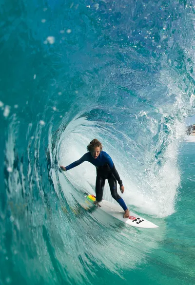A surfer rides a large, clear blue wave, surrounded by ocean spray, with a sandy beach and cliffs in the background under a clear sky.