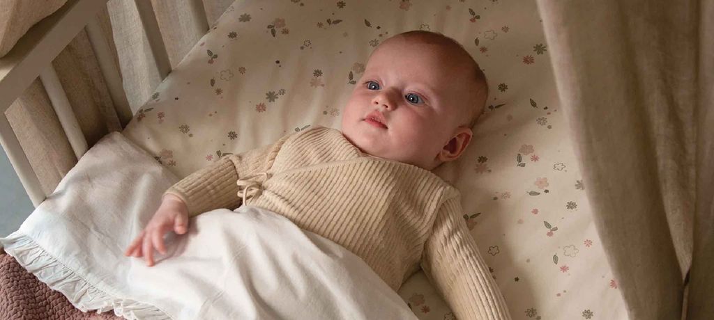 Baby in a beige sweater lies in a crib on a floral-patterned sheet, partially covered by a white blanket.