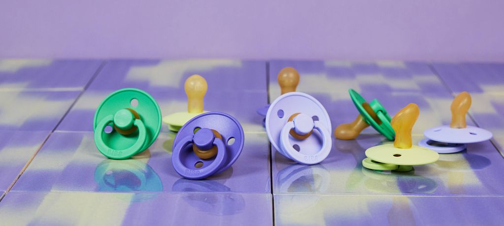 Colorful pacifiers in green, purple, and yellow arranged on a reflective, purple-tiled surface.