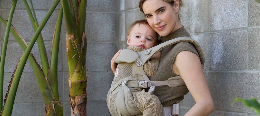 Woman holding a baby in a beige carrier, standing outdoors near a palm tree against a concrete wall.