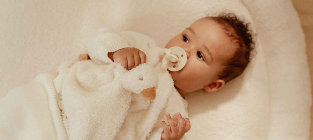 Baby lying on a soft blanket, holding a plush toy, with a pacifier in mouth, wearing a white sweater.
