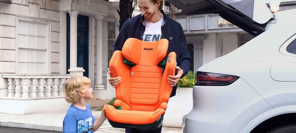 Man smiling, holding an orange Recaro child car seat near a white car, with a boy looking on.