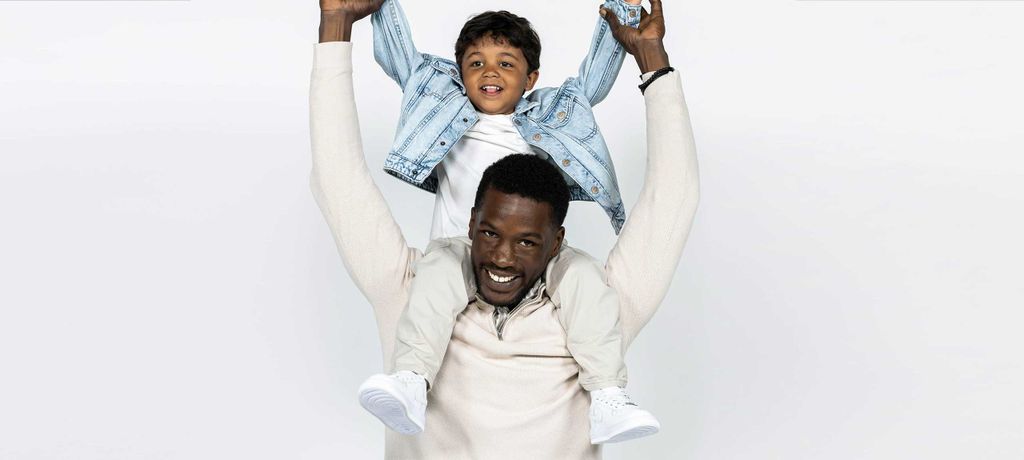 Man smiling with a child sitting on his shoulders, both wearing light-colored clothes, against a plain white background.