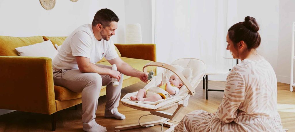 Parents smile at their baby in a bouncer, engaging with toys, in a cozy living room with a mustard sofa.