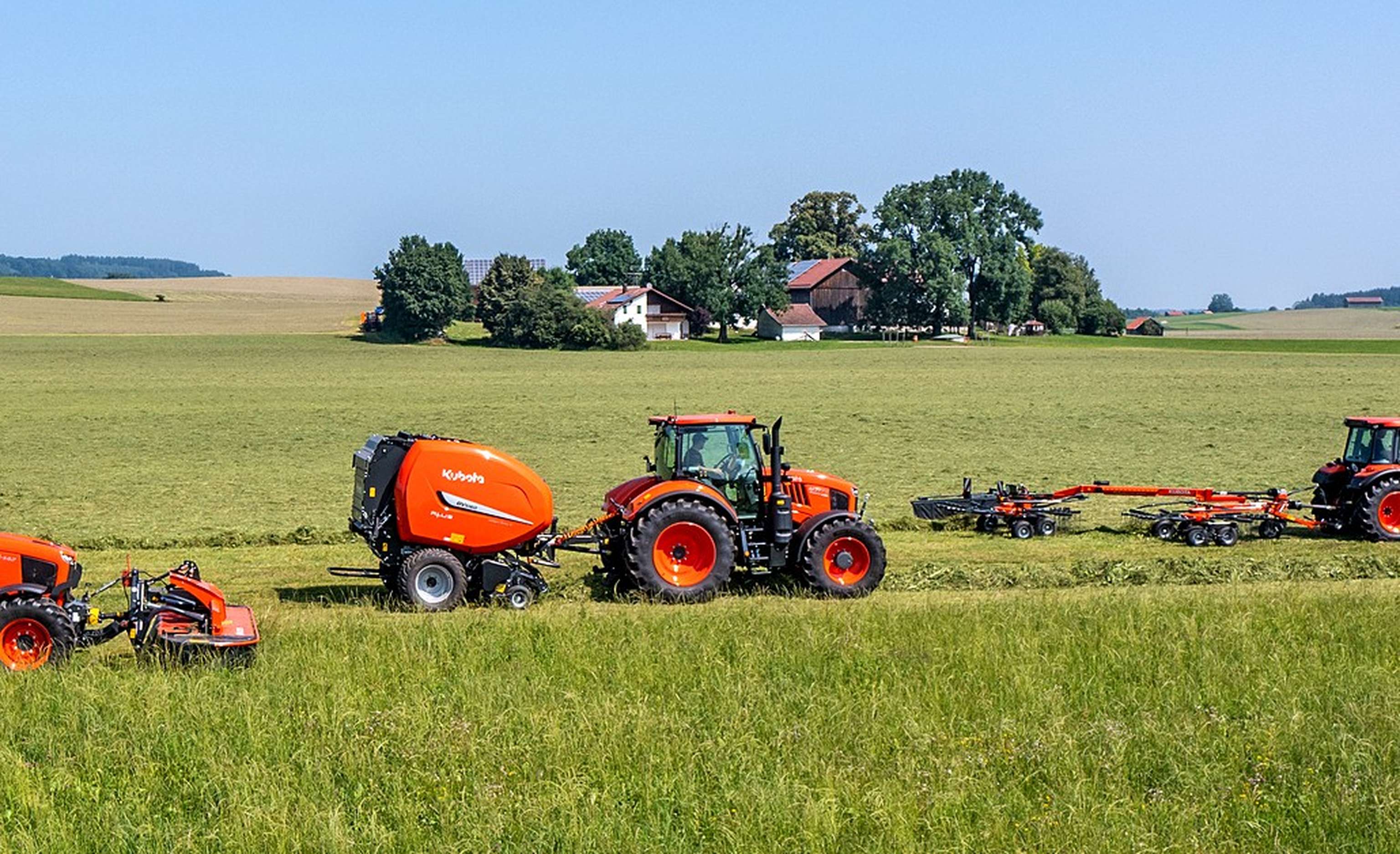 Tractors and machinery work in a green field with farm buildings and trees in the background under a clear blue sky.