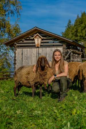 Junge Frau hockt lächelnd neben einem braunen Schaf auf einer grünen Wiese vor einem rustikalen Holzstall bei blauem Himmel.