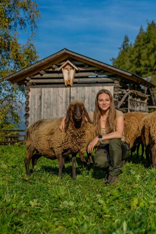 Junge Frau hockt lächelnd neben einem braunen Schaf auf einer grünen Wiese vor einem rustikalen Holzstall bei blauem Himmel.