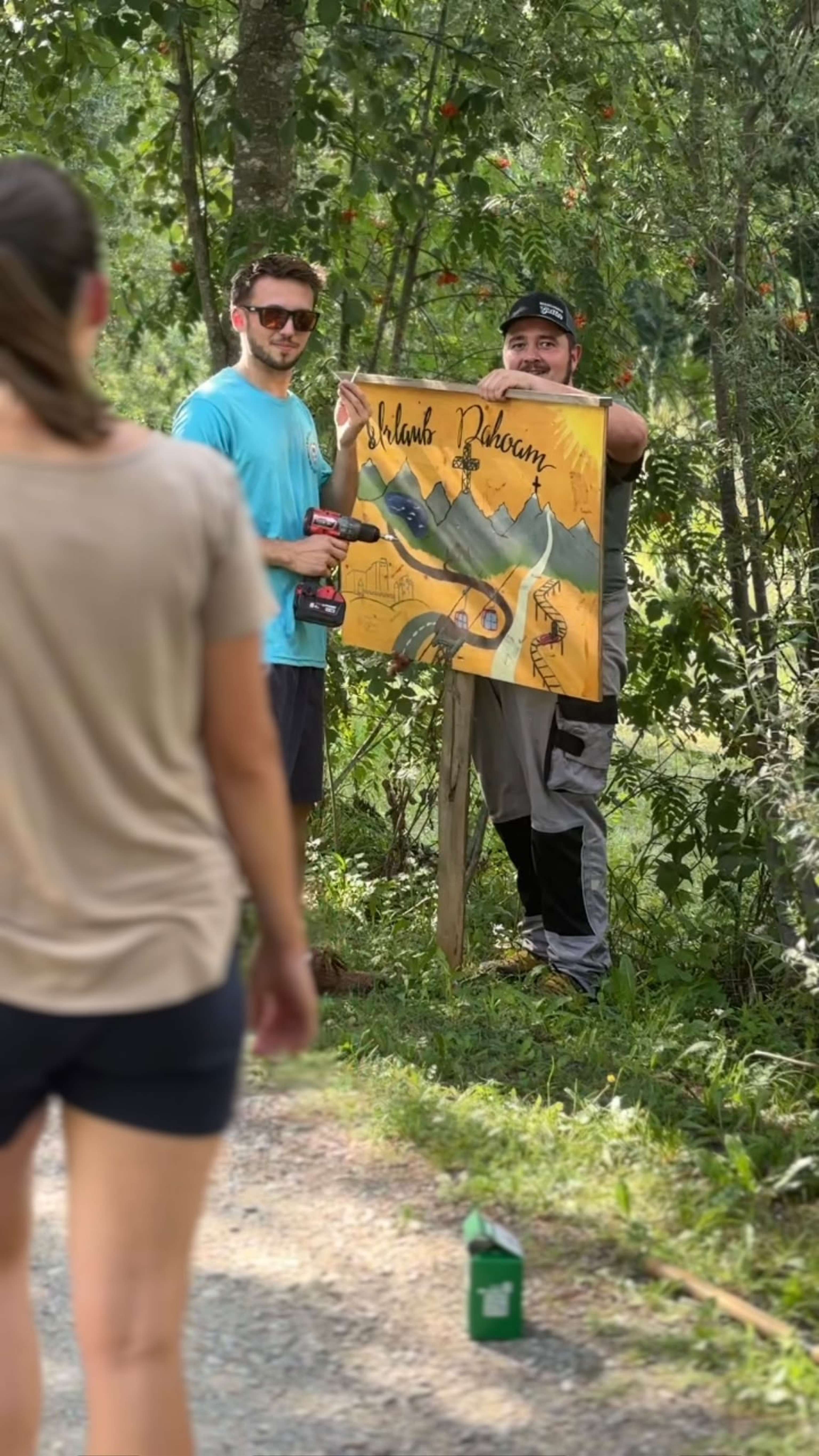 Zwei Männer im Wald montieren ein gelbes handgemaltes Schild mit Berglandschaft, einer hält einen Akkuschrauber.