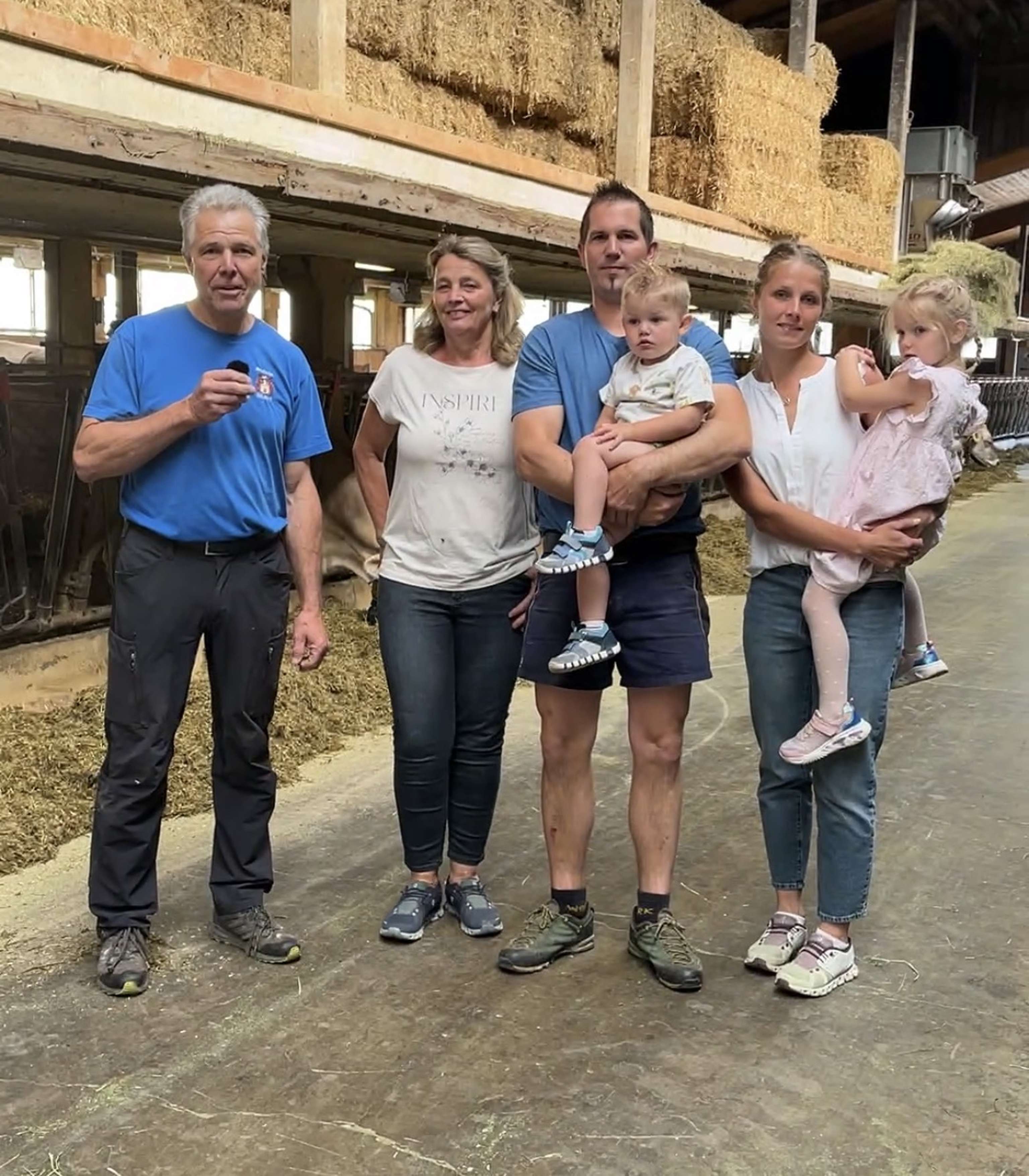 Fünf Erwachsene und zwei Kinder stehen in einem Stall mit Strohballen im Hintergrund. Ein Mann hält ein Mikrofon.