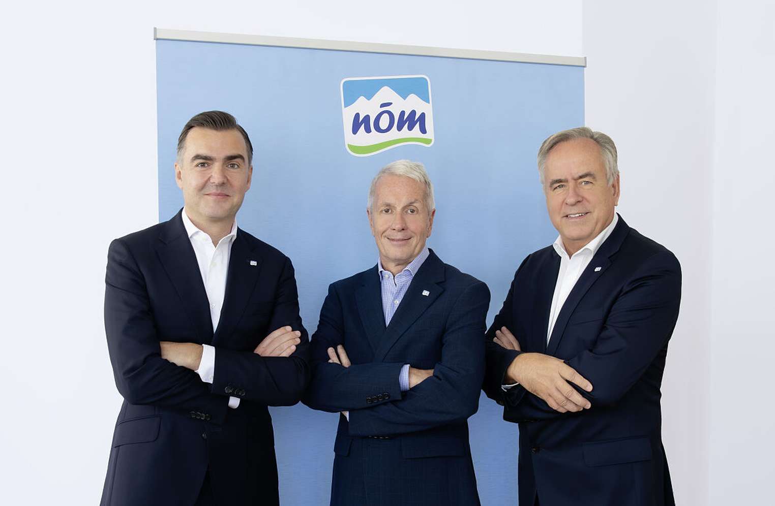 Drei Männer in Anzügen posieren mit verschränkten Armen vor einem blauen NÖM-Marken-Banner.