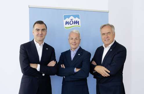 Drei Männer in Anzügen posieren mit verschränkten Armen vor einem blauen NÖM-Marken-Banner.