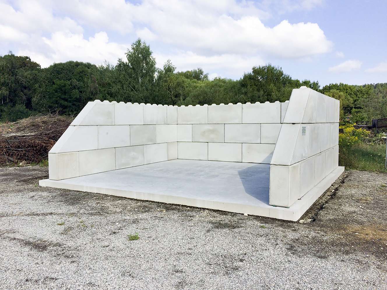 U-förmige Betonmauer aus gestapelten Betonblöcken auf einer Betonplatte im Freien, umgeben von Bäumen und bewölktem Himmel.