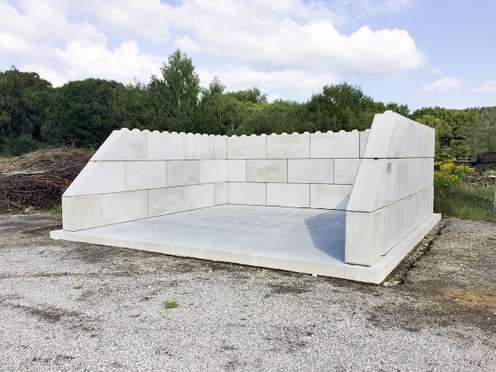 U-förmige Betonmauer aus gestapelten Betonblöcken auf einer Betonplatte im Freien, umgeben von Bäumen und bewölktem Himmel.