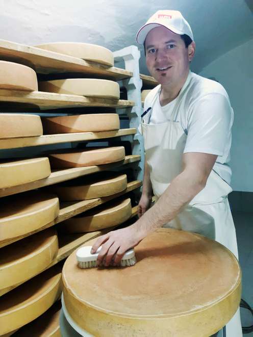 Ein Mann in weißer Kleidung bürstet einen großen Käselaib in einem Lagerraum mit mehreren Regalen voller Käse.