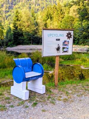 Blauer Sessel aus recycelten Fässern auf weißem Holzsockel neben einem Upcycling-Infotafel, umgeben von Natur und einem Teich.