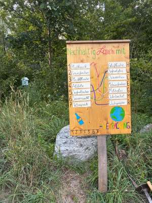 Handgemaltes Holzschild im Freien mit Aufschrift „Nachhaltig Leben mit Verpackung", zeigt Wegwerf- vs. Mehrwegverpackungen in der Natur.
