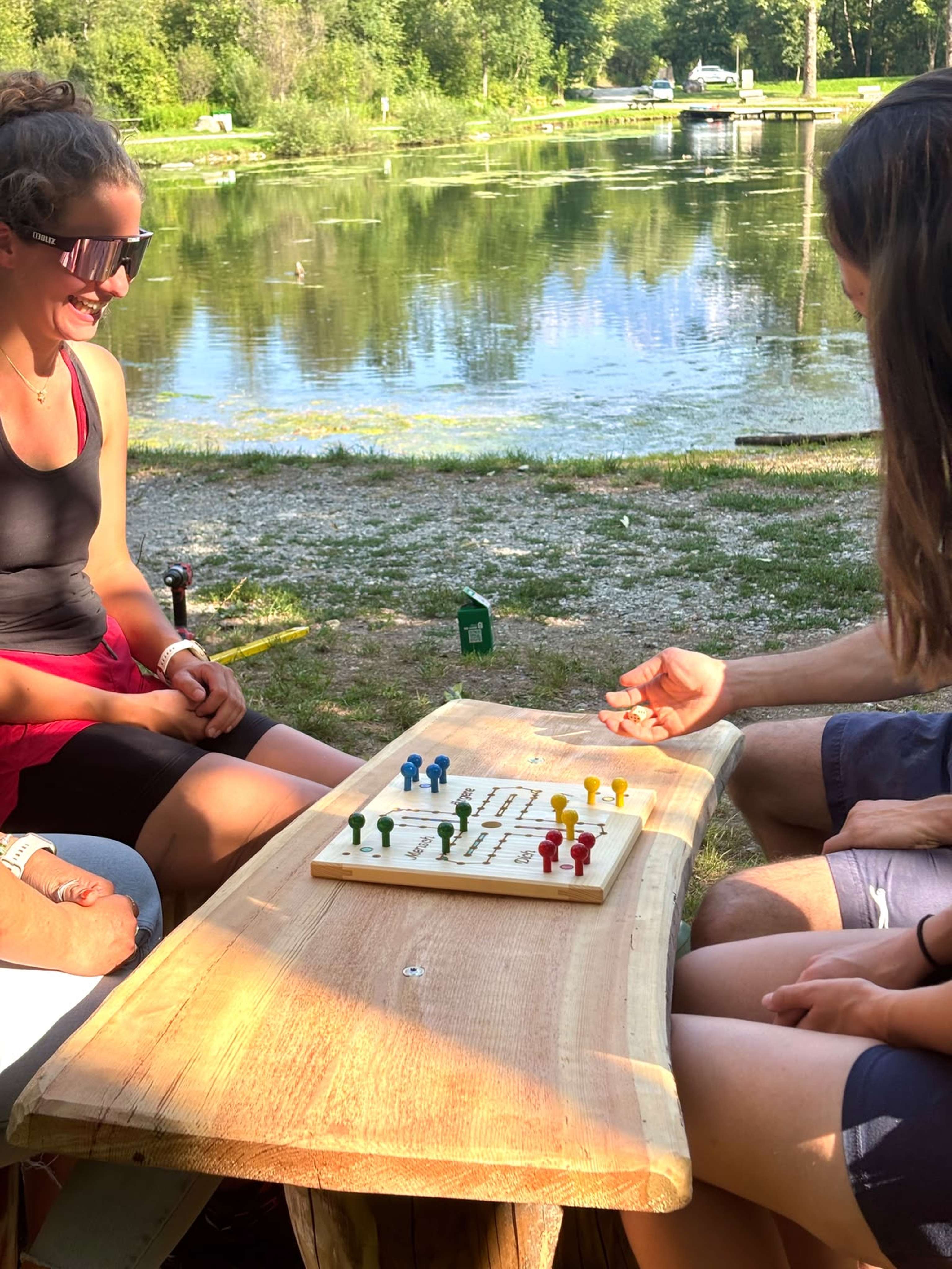 Zwei Frauen spielen Mensch-ärgere-dich-nicht an einem Holztisch im Freien, im Hintergrund ein ruhiger Teich mit Bäumen.