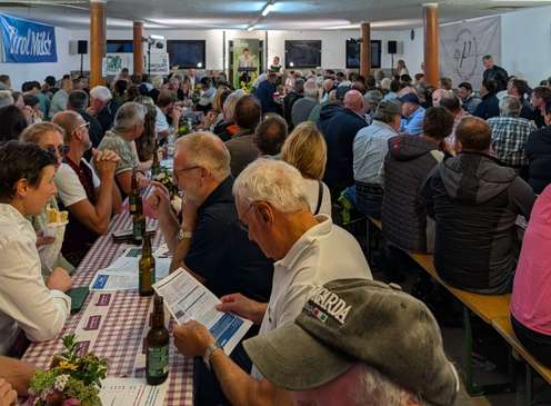 Vollbesetzter Veranstaltungssaal mit vielen Menschen an langen Tischen, Tirol Milch Banner, Redner vorne, Bierflaschen auf karierten Tischdecken.