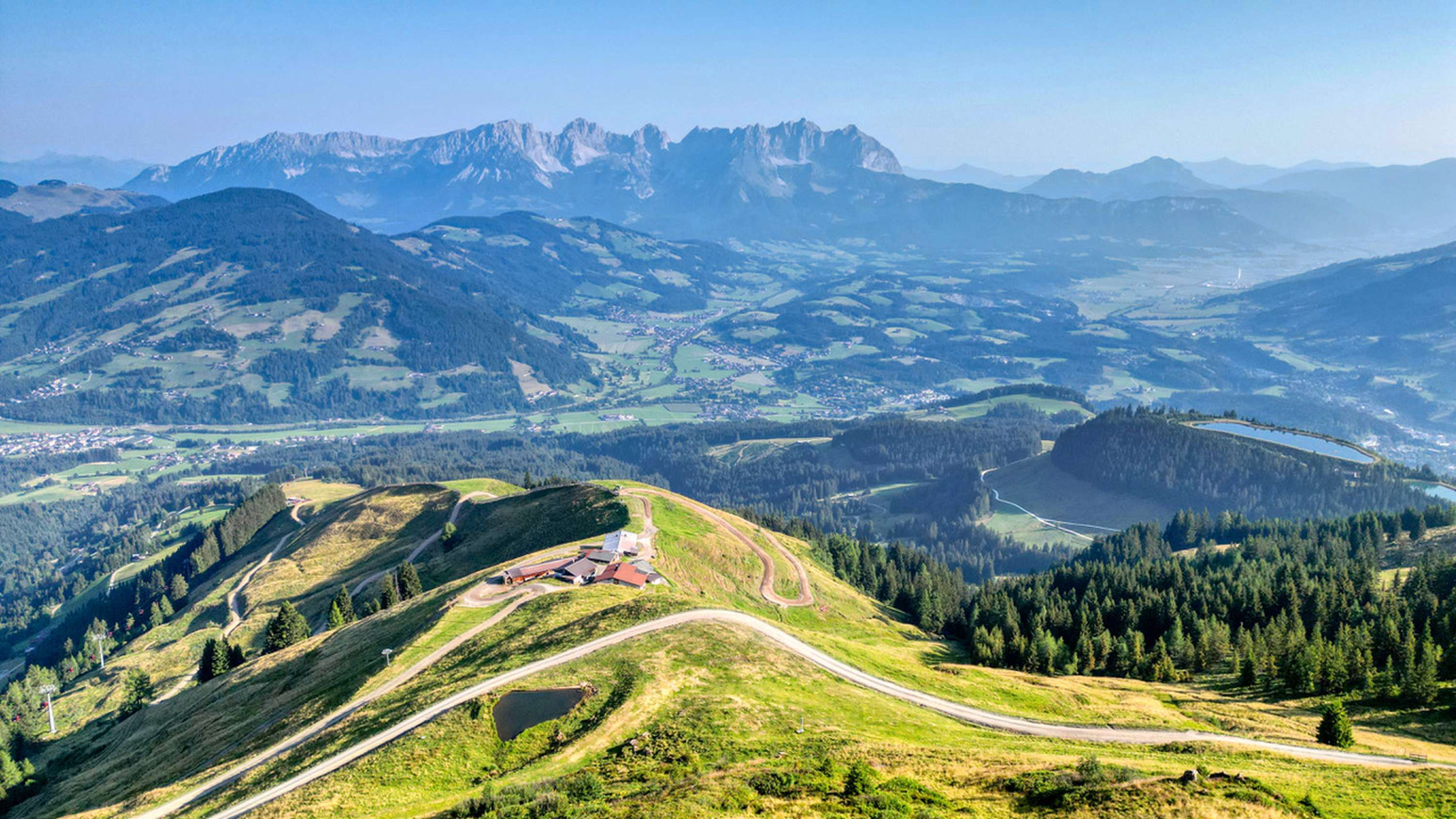 Luftpanorama einer Almstation in den Alpen mit geschwungenen Wegen, grünen Hängen, Tälern und dem Kaisergebirge im Hintergrund.