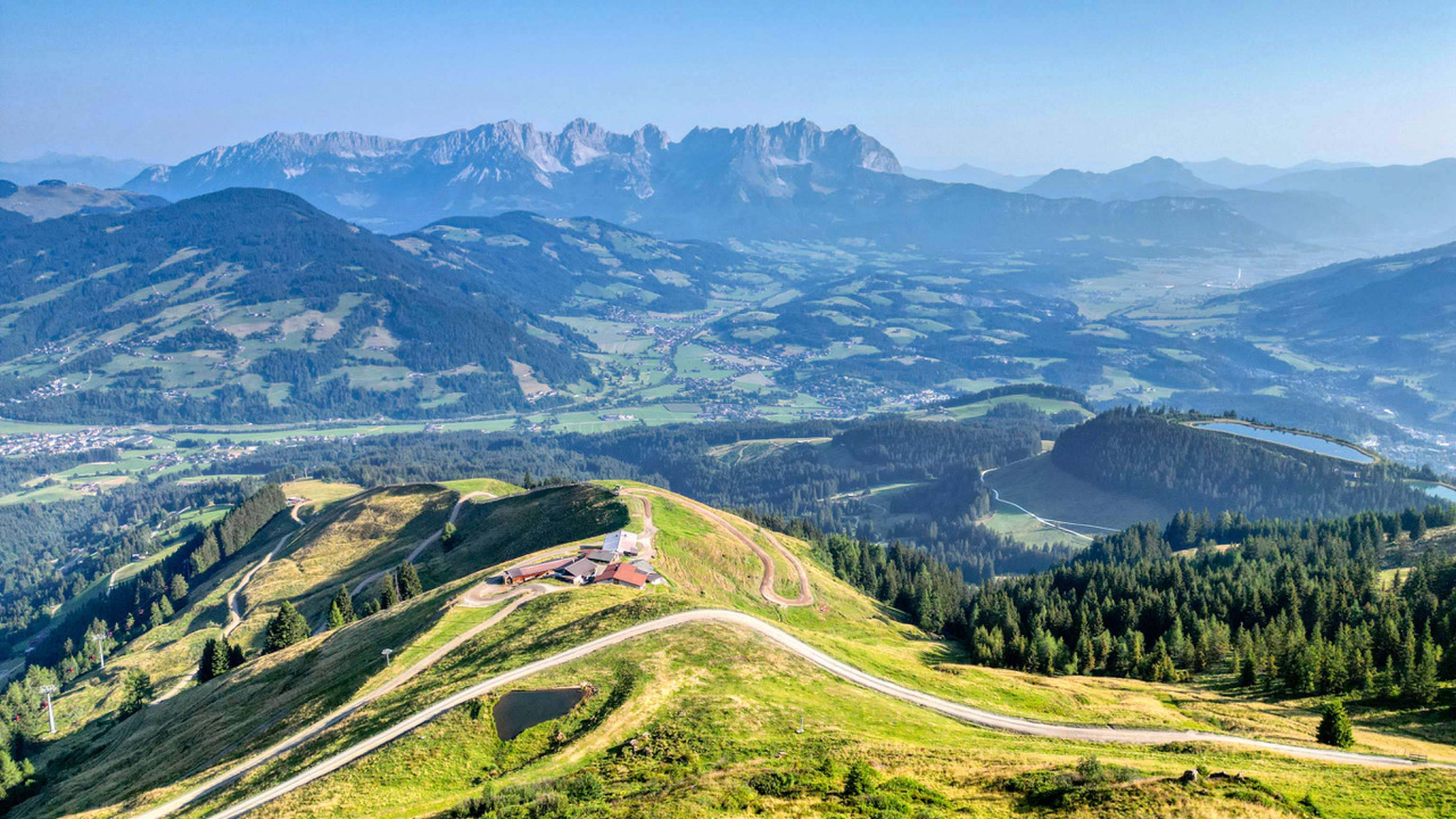 Luftpanorama einer Almstation in den Alpen mit geschwungenen Wegen, grünen Hängen, Tälern und dem Kaisergebirge im Hintergrund.