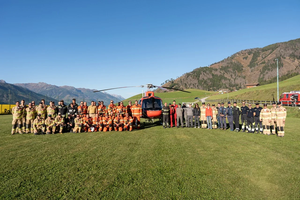 A large group of rescue personnel in various uniforms stand in front of a red helicopter on a grassy field, with mountains in the background.