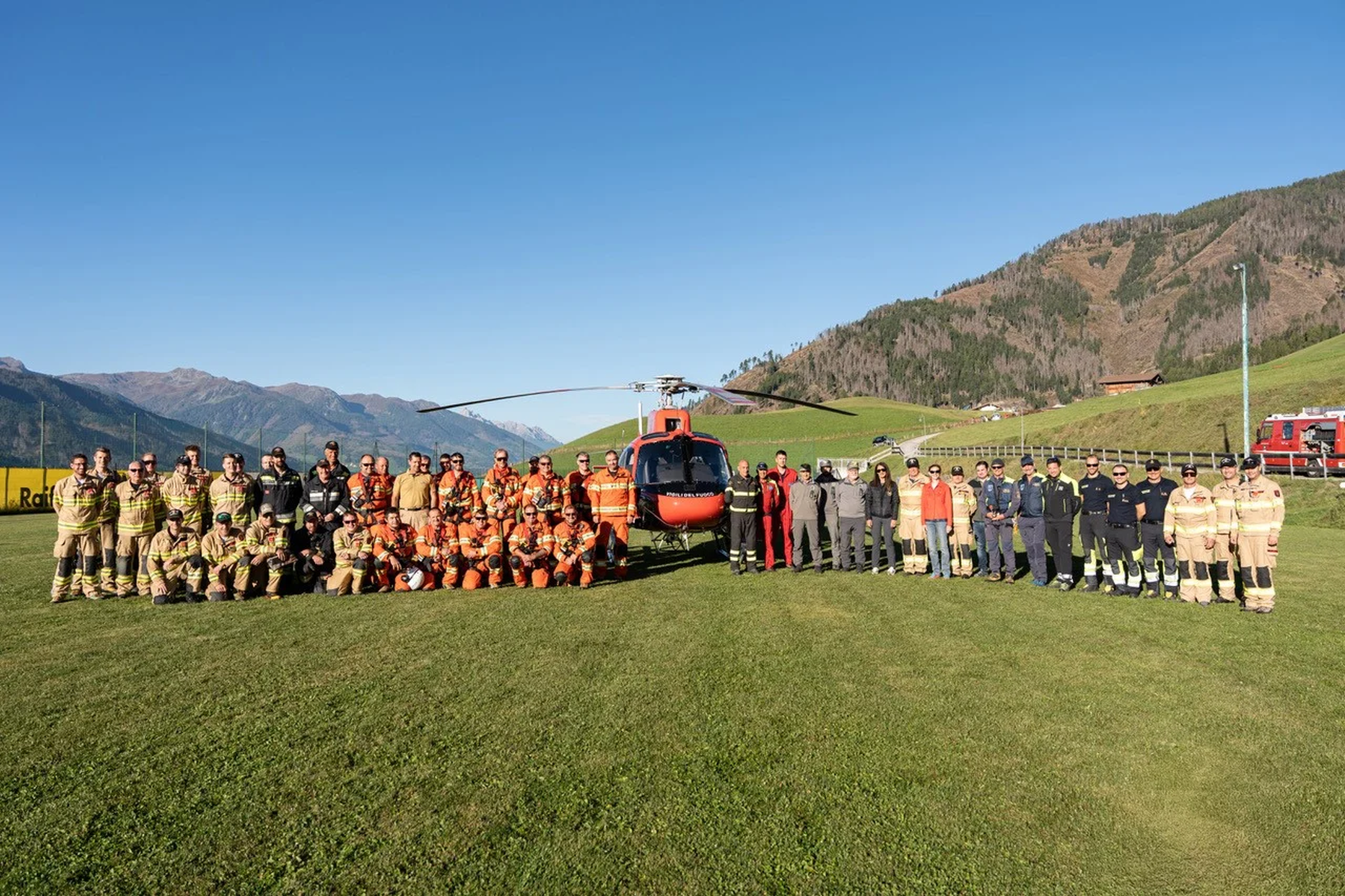 A large group of rescue personnel in various uniforms stand in front of a red helicopter on a grassy field, with mountains in the background.