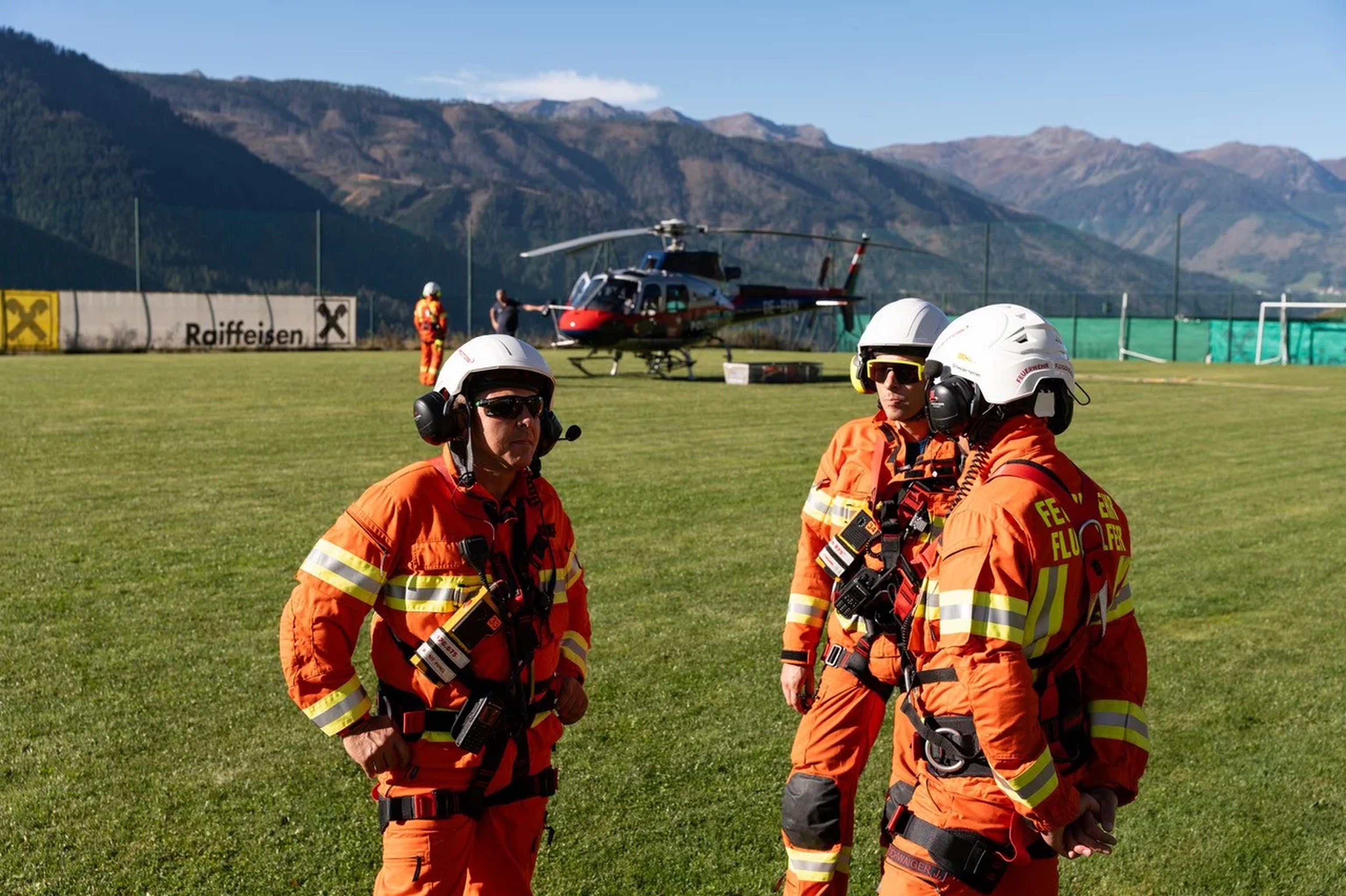 Three rescue workers in orange uniforms and helmets stand on a grassy field with a helicopter and mountains in the background.