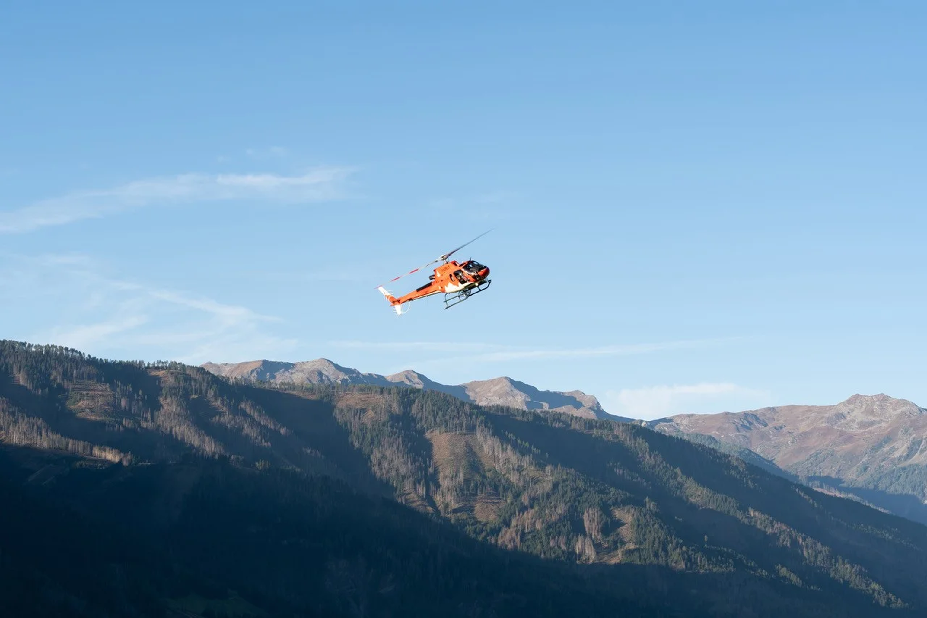 An orange helicopter flies over a mountainous landscape under a clear blue sky.