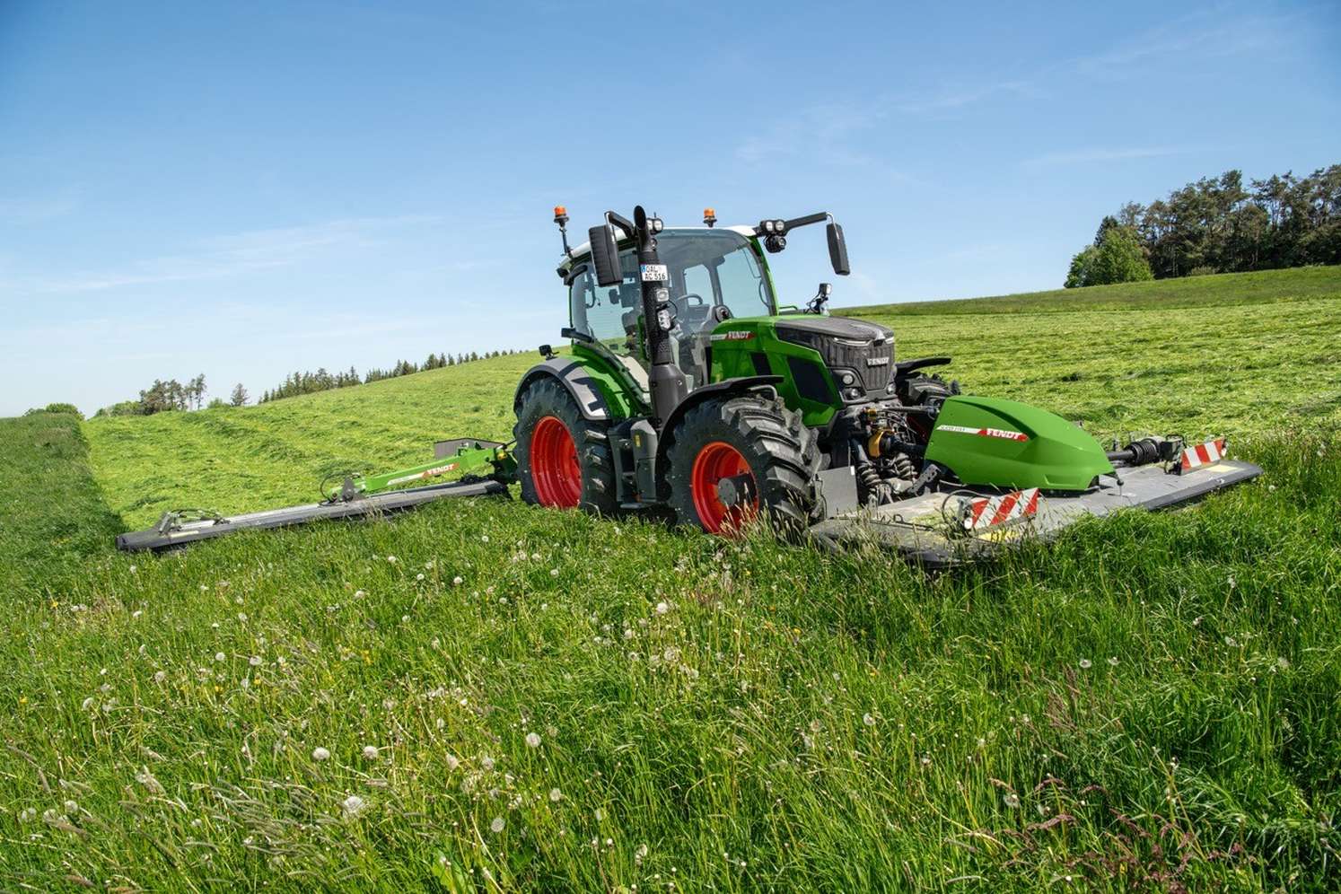 Grüner Fendt-Traktor mit roten Felgen mäht eine grüne Wiese mit breitem Mähwerk bei blauem Himmel.