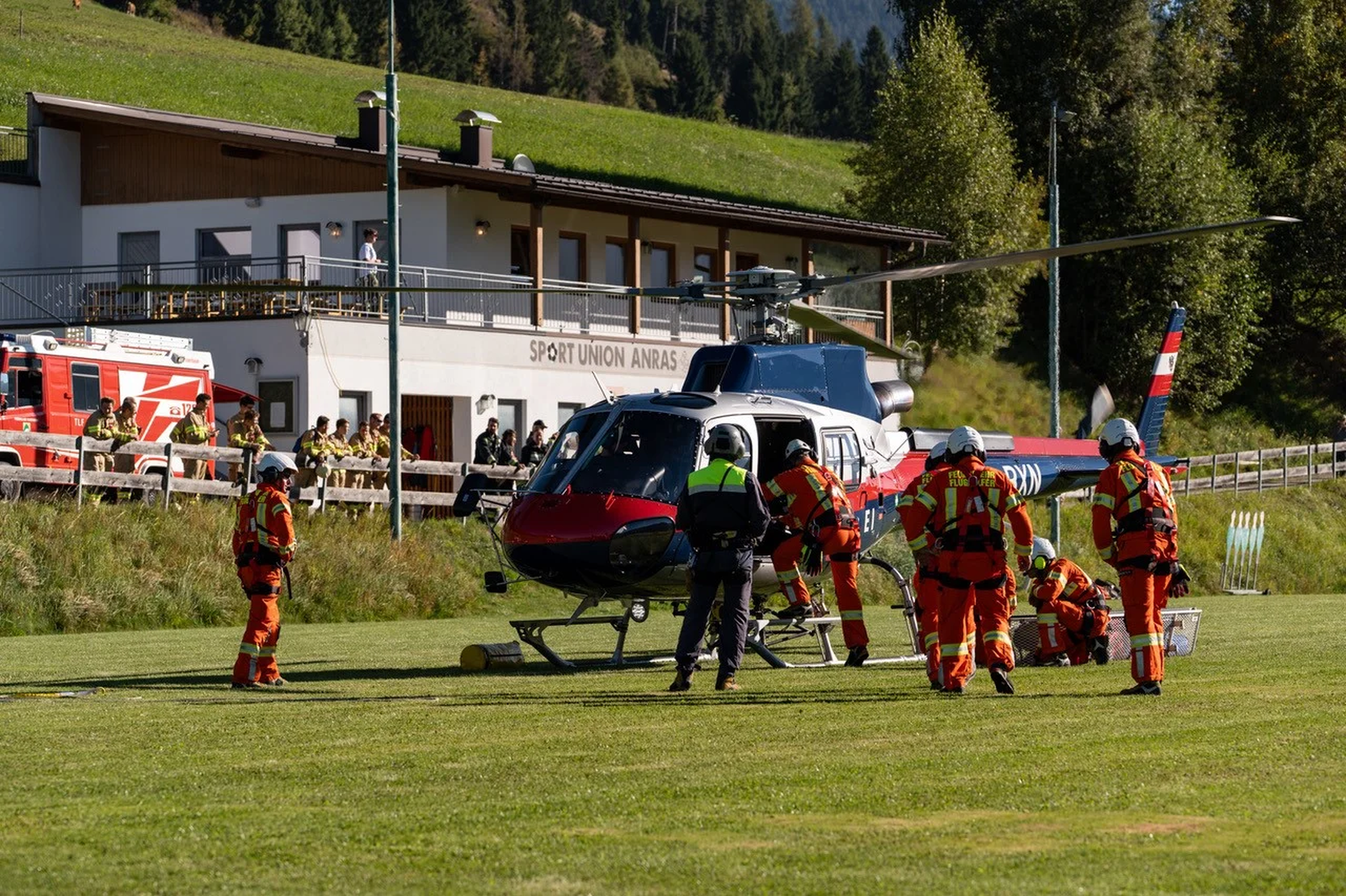 Rescue workers in orange uniforms attend to a helicopter on a grassy field near a building, with emergency vehicles in the background.
