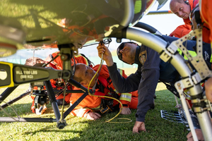 Rescue team members in orange suits inspect the underside of a helicopter on grass, with one member using a flashlight.