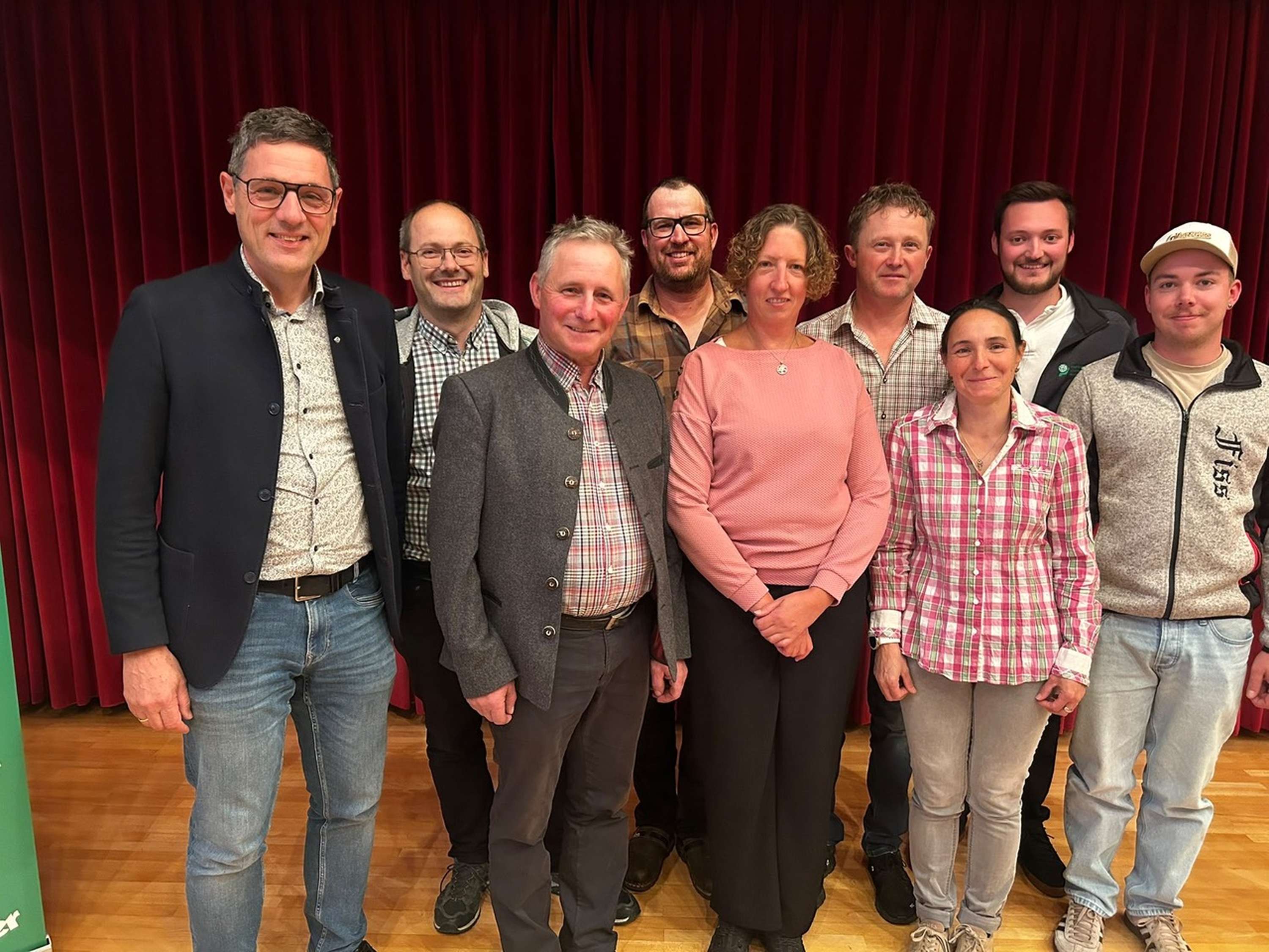 Gruppe von neun Personen, Männer und Frauen, posieren lächelnd vor einem roten Samtvorhang in einem Veranstaltungssaal.