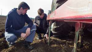Zwei junge Männer untersuchen gemeinsam den Boden neben einem Traktor auf einem Feld.