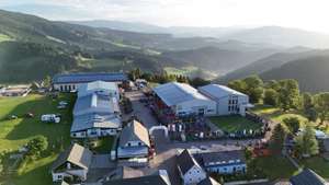 Luftaufnahme des SOMA-Firmengeländes mit Lagerhallen, Landmaschinen und Solaranlagen, umgeben von grüner Mittelgebirgslandschaft.