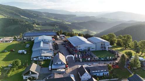 Luftaufnahme des SOMA-Firmengeländes mit Lagerhallen, Landmaschinen und Solaranlagen, umgeben von grüner Mittelgebirgslandschaft.