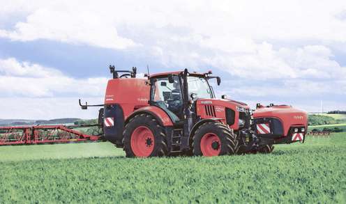Red tractor with a sprayer attachment driving through a lush green field under a partly cloudy sky.